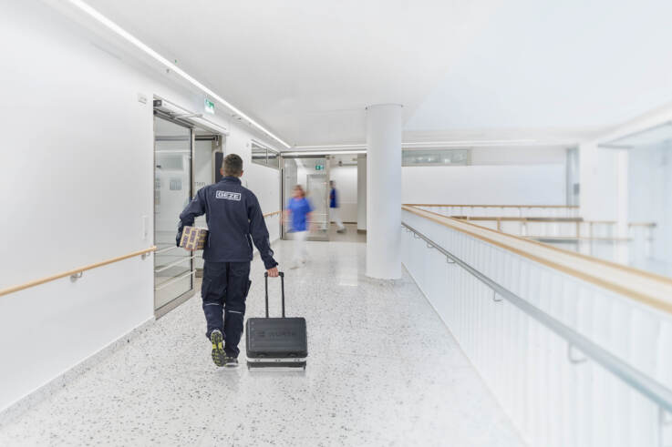 A GEZE service technician walks down a hospital corridor carrying a toolbox and a door closer
