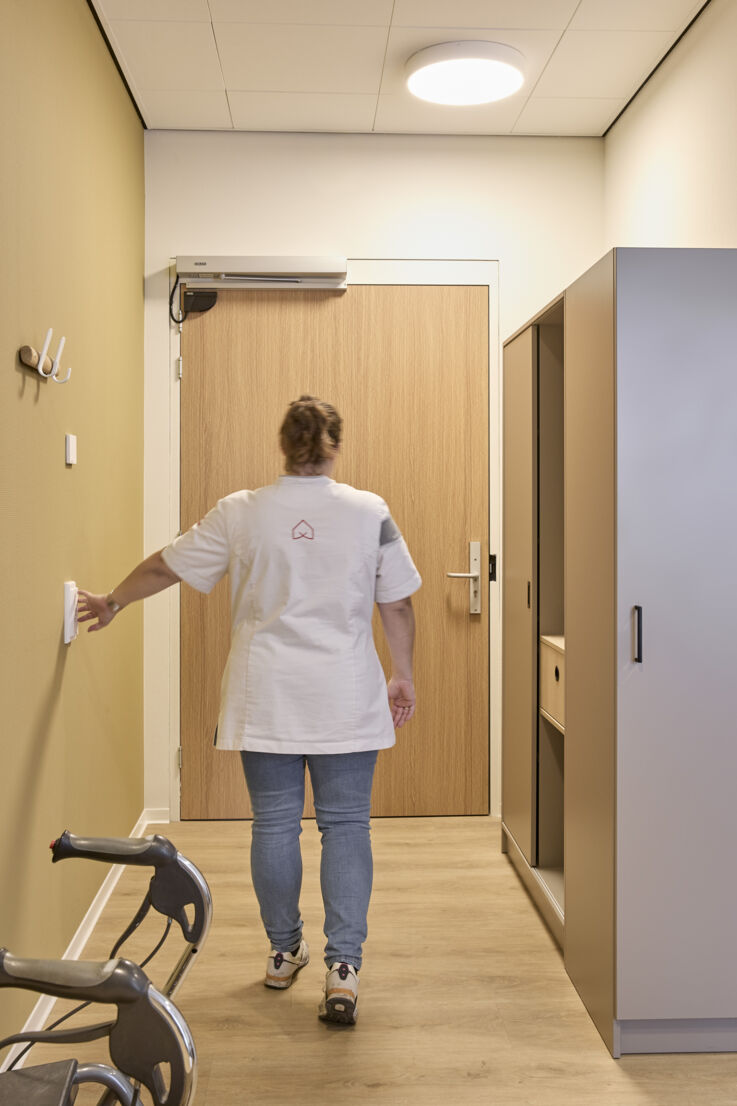 A nurse walks towards a door and opens it using a manual switch on the wall.