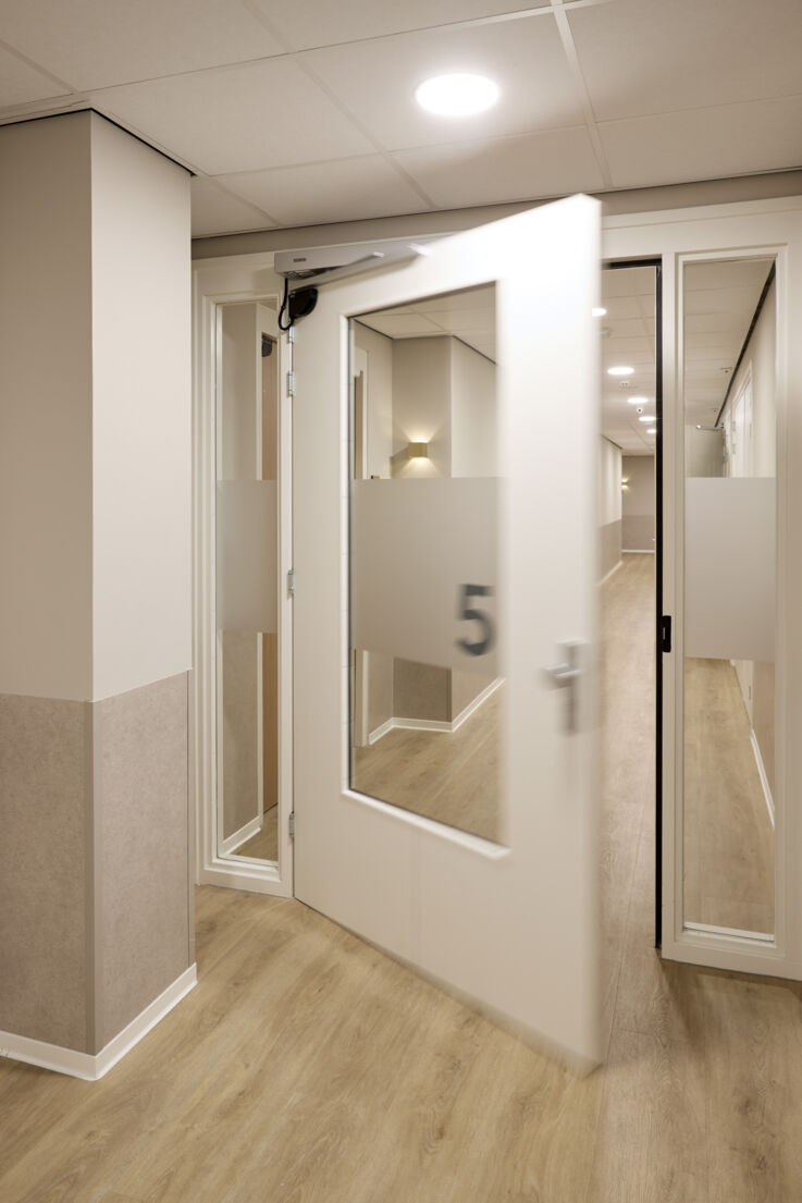 A half-open glass door with automatic drive units in a modern nursing home corridor.